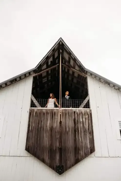 A bride and groom stand in the open, dark hayloft of a white barn, looking in opposite directions.