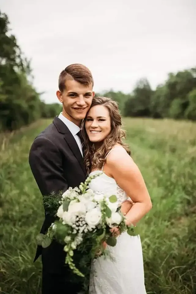 A couple in wedding attire poses affectionately in a grassy field while holding a bouquet of white flowers.