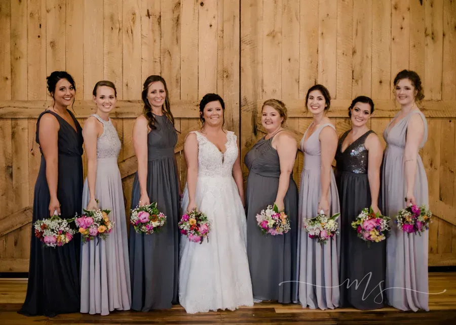 A bride in a white wedding gown stands with bridesmaids in alternating grey and lavender dresses before wood doors.