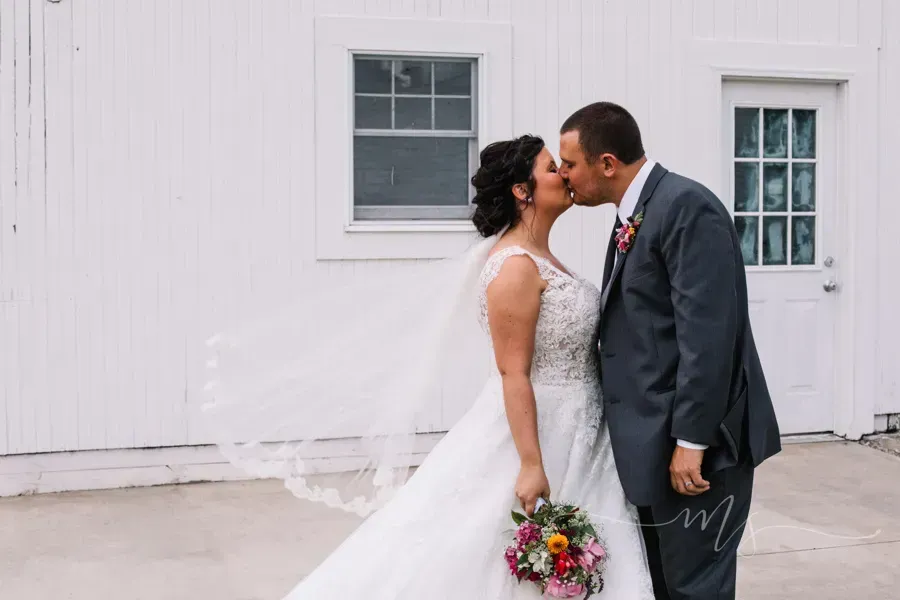 A wedding couple kisses while standing in front of a white wall with a window and a door, holding a colorful bouquet.