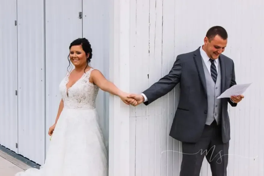 A bride and groom holding hands around a corner of a white wall, with the groom reading from a piece of paper.