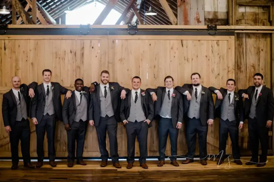 Nine groomsmen in dark suits and gray vests stand in a row smiling in front of rustic wooden barn doors.