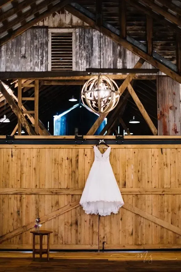 A white wedding dress hangs centered on a large wooden barn door beneath a spherical chandelier in a rustic venue.