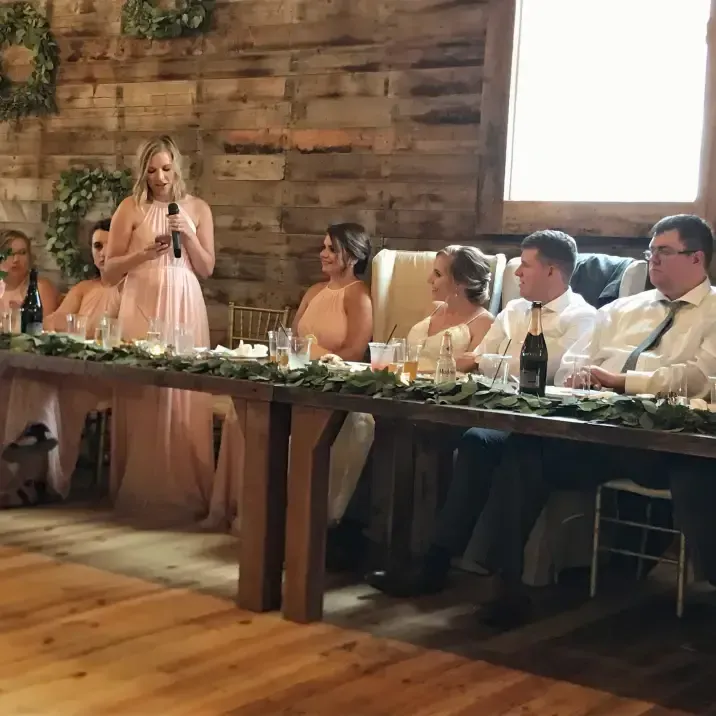 A person speaks into a microphone at a wedding head table while others seated in formal wear listen in a rustic venue.