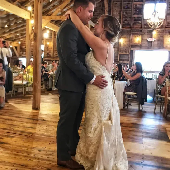 A couple dances at their wedding reception inside a rustic, wood-paneled barn with warm lighting and guests in the back.
