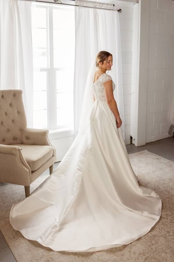 A person in a white wedding gown with a lace bodice and long train stands in a sunlit room near a beige armchair.