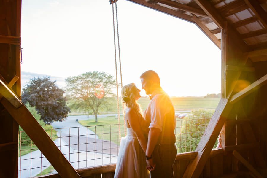 A couple in wedding attire stand holding hands, framed by the rustic timber structure of a barn, backlit by a sunset.