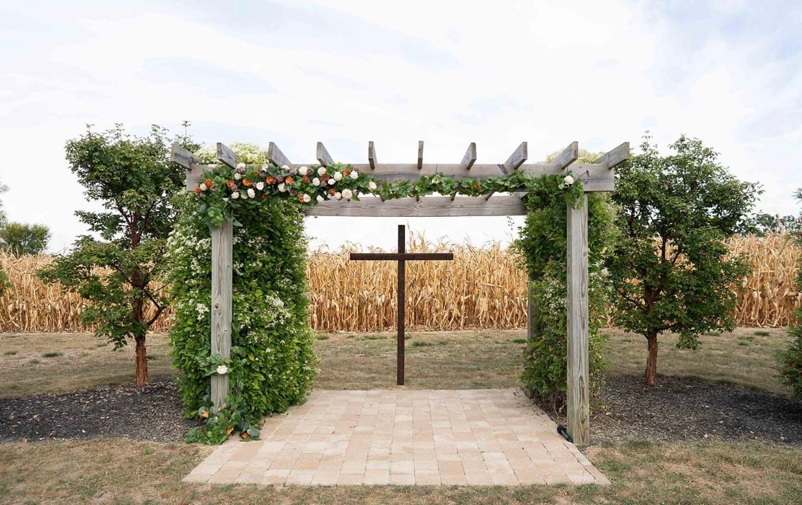 A wooden pergola draped in greenery and flowers stands over a stone patio with a large cross, set before a cornfield.