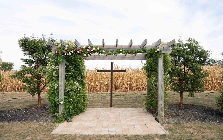 A wooden arbor with climbing vines and flowers stands over a stone patio with a metal cross, set before a cornfield.
