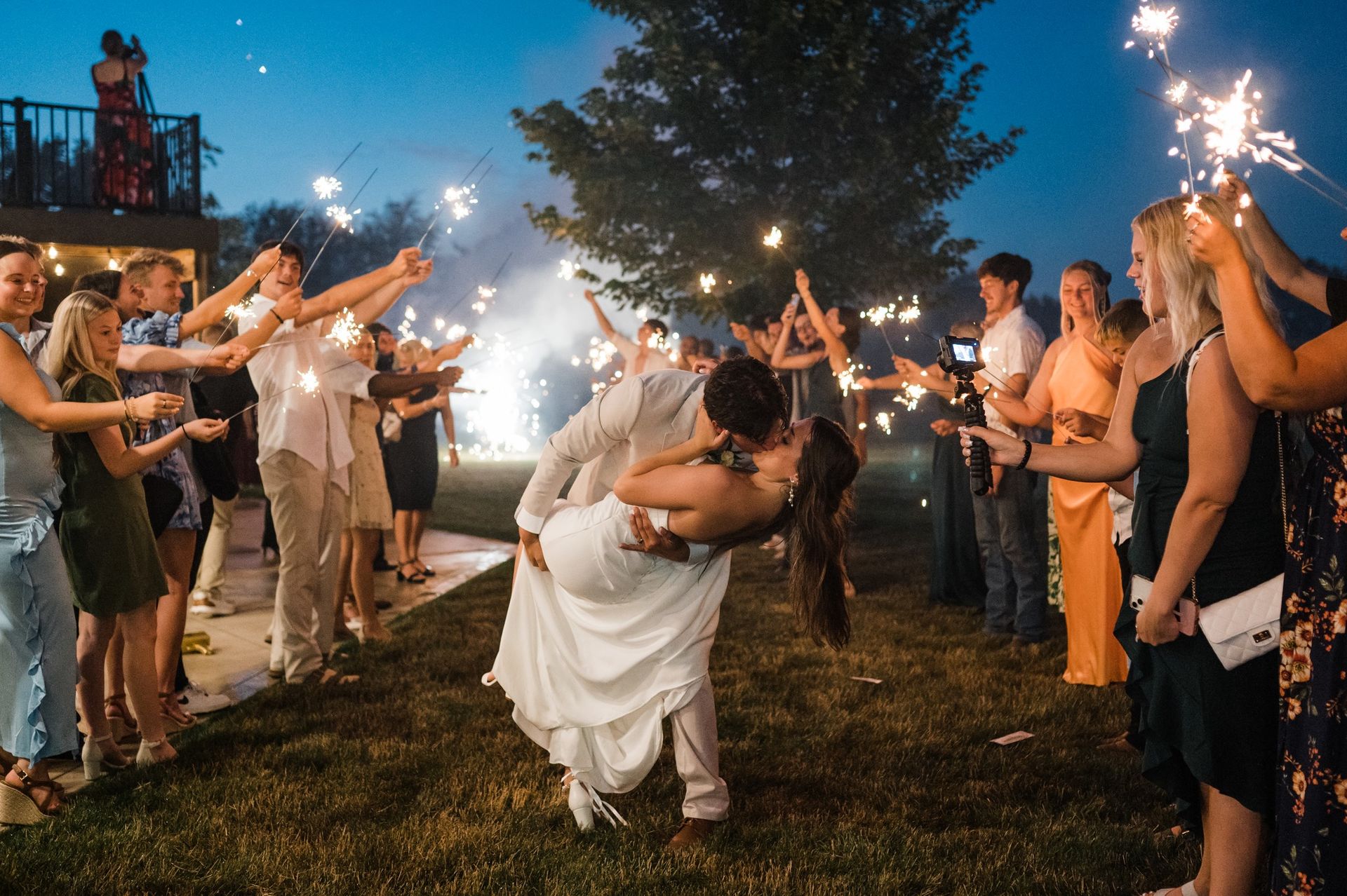 A couple kisses in a dip while surrounded by wedding guests holding lit sparklers at dusk.