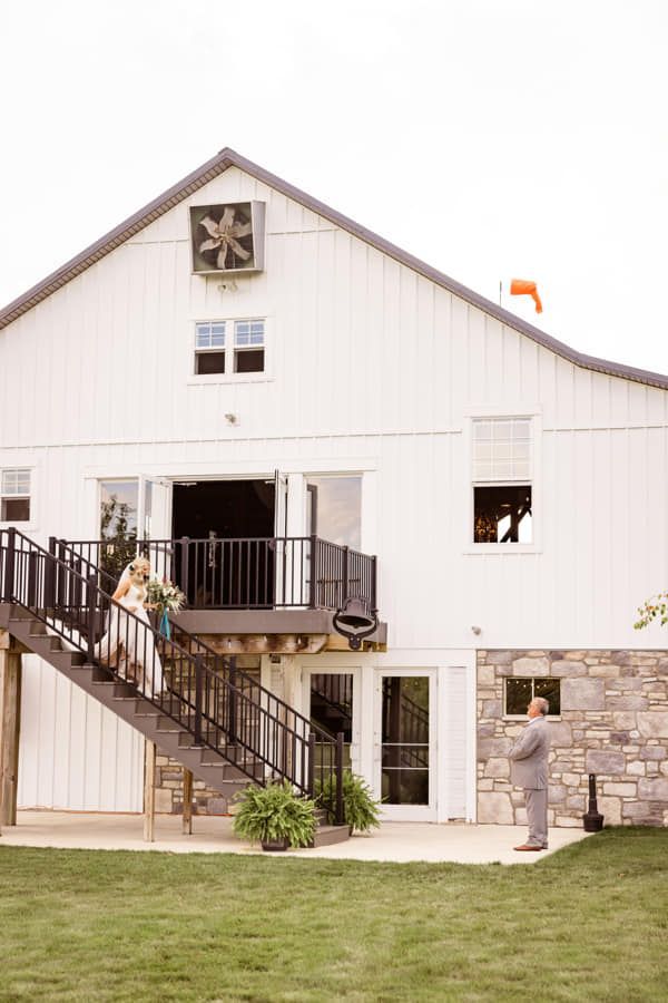 A person in a wedding dress descends stairs toward a person in a suit, outside a white barn venue.