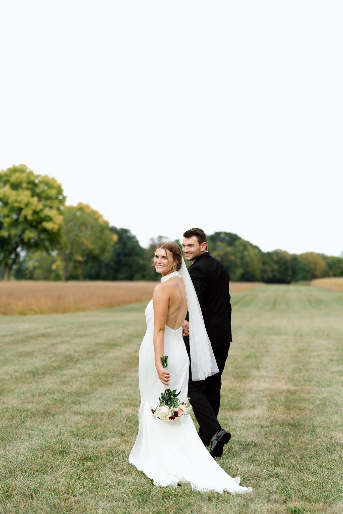 A wedding couple walks through a grassy field, looking back toward the camera, the bride holding a small bouquet.