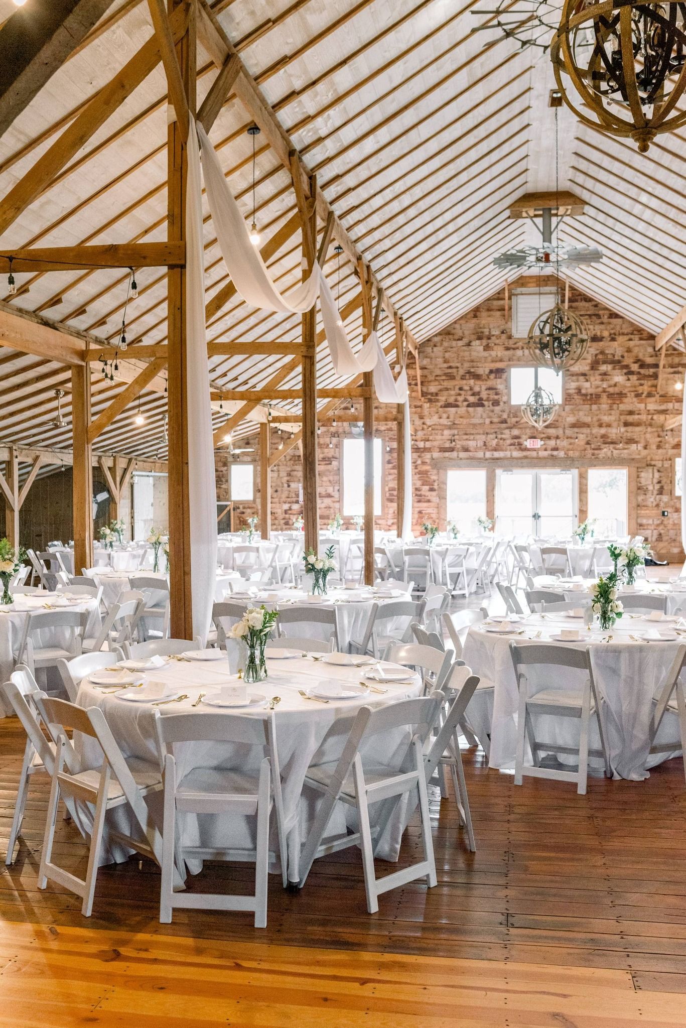 Round tables with white linens and chairs set up for a wedding reception in a rustic barn with exposed wood rafters.