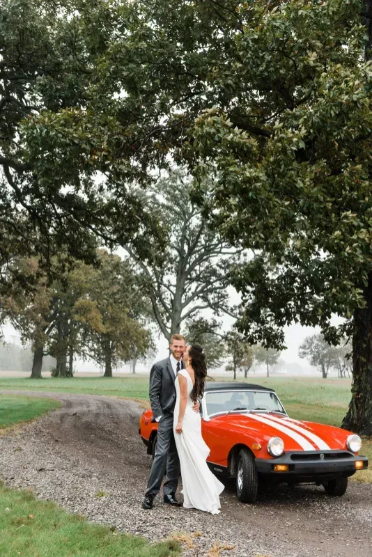 A couple in wedding attire embraces beside a bright orange sports car with white racing stripes on a gravel path.