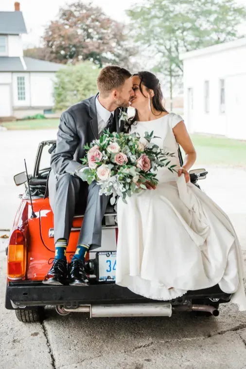 A bride and groom in wedding attire kiss while sitting on the back of an orange vintage convertible car.