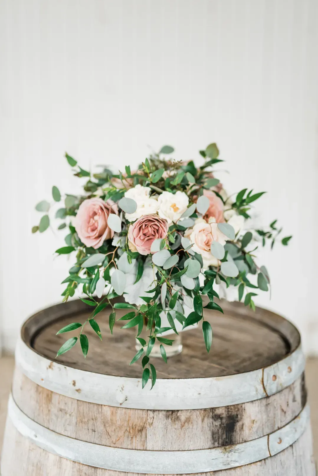 A bouquet of dusty pink and white roses with eucalyptus greenery, displayed in a glass vase atop a rustic wooden barrel.