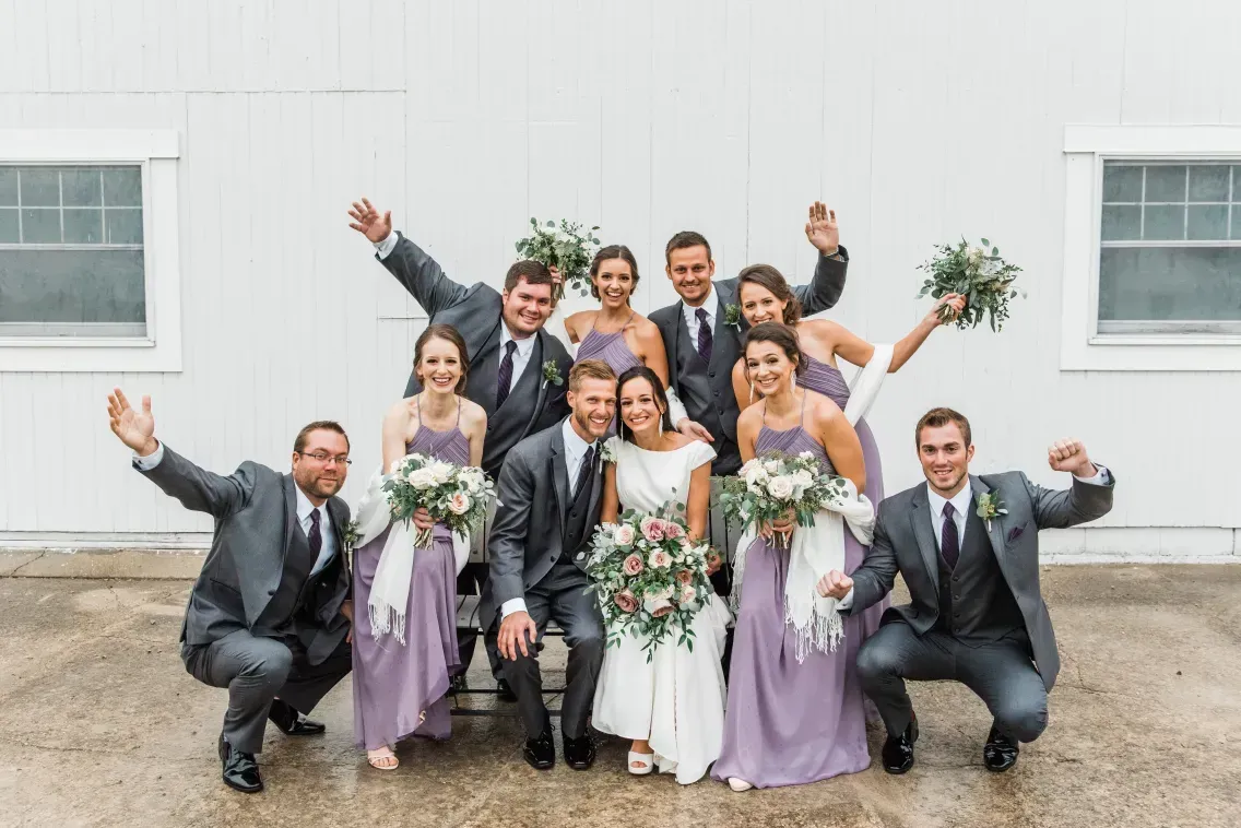 A wedding party in gray suits and lavender dresses poses for a group photo in front of a white barn.