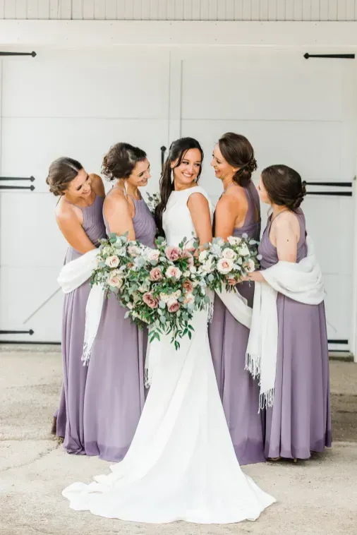 A bride in a white gown and four bridesmaids in dusty purple dresses stand in a circle holding bouquets before white doors.
