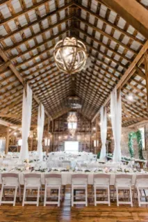 An interior view of a rustic wooden barn set up for a wedding reception with long tables, white chairs, and chandeliers.