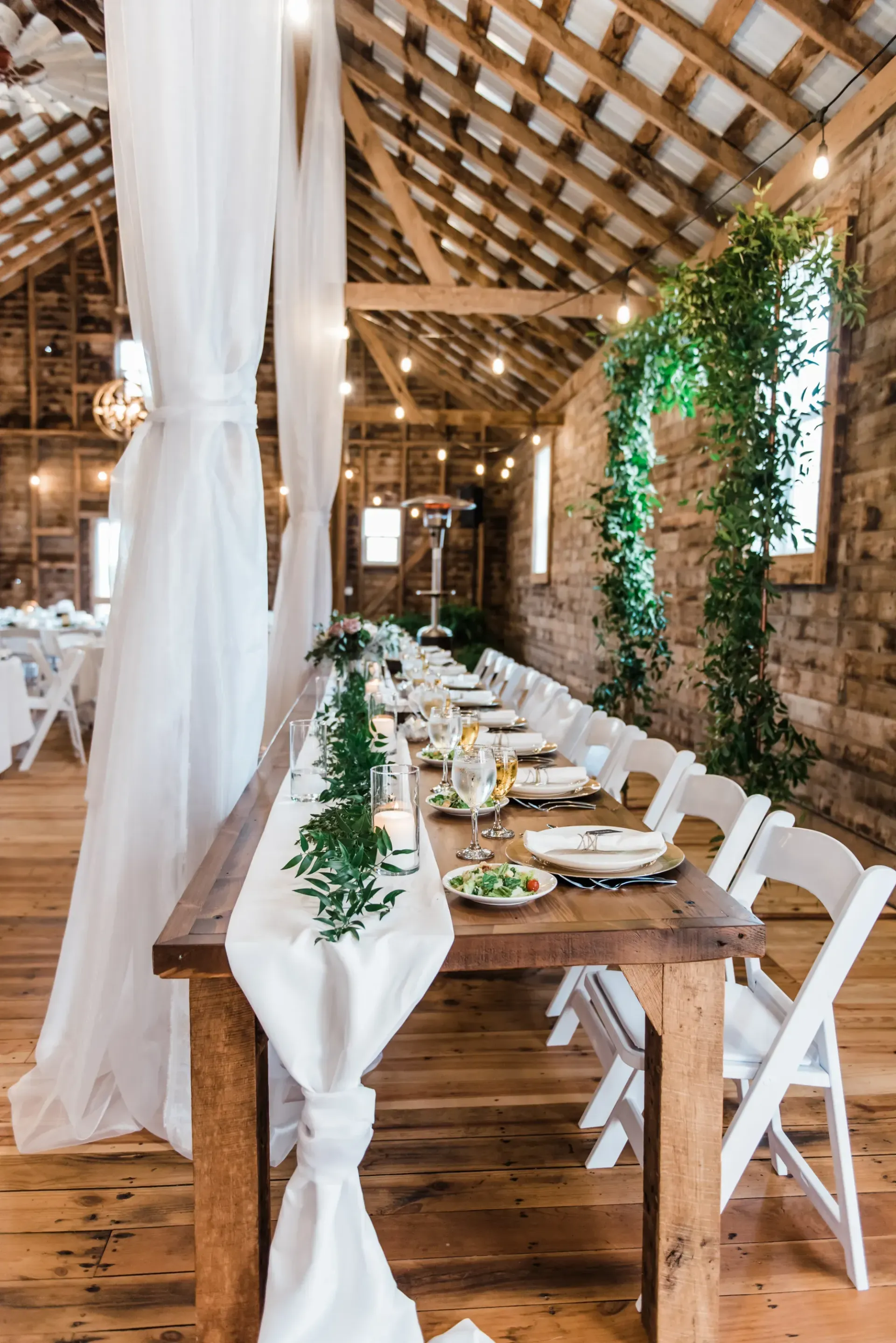 A long, wooden dining table decorated with white fabric and greenery inside a rustic barn with string lights.