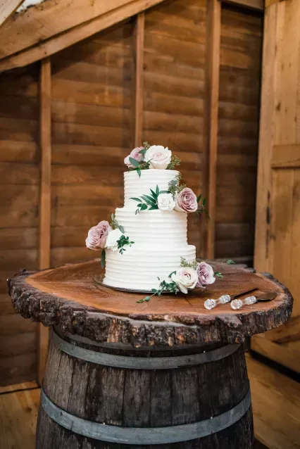 A three-tiered white wedding cake with floral accents, displayed on a rustic wood-slice table set atop a wine barrel.