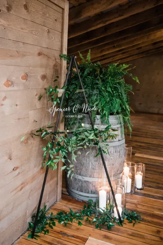 Wedding welcome sign on a black triangular frame with greenery, set beside a wooden wine barrel and candles on floor.