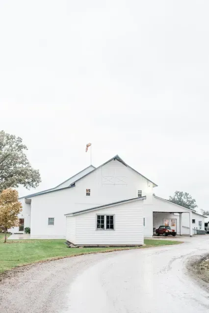 A white barn with a metal roof sits beside a paved driveway on a cloudy day, with a windsock on its roof.