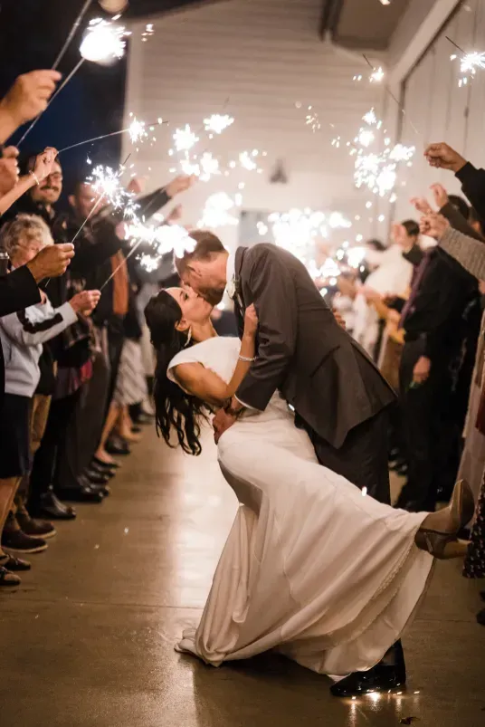 A couple in formal wedding attire shares a dip and a kiss while surrounded by guests holding lit sparklers at night.