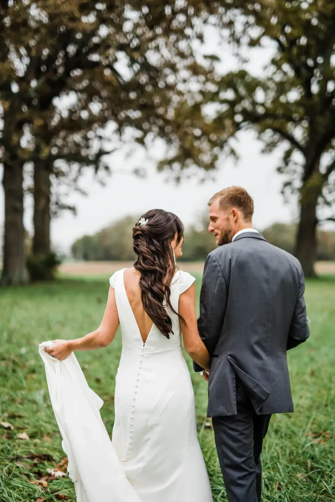 A couple in wedding attire walking hand-in-hand through a grassy field with trees in the background.