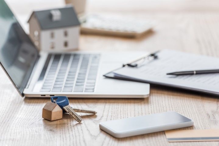 Wooden desk with a laptop, house key, smartphone, and clipboard