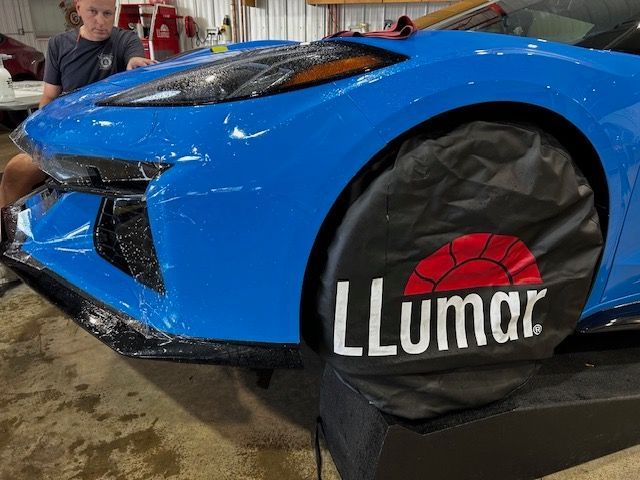 A man is sitting in front of a blue car with a llumar tire cover on it.
