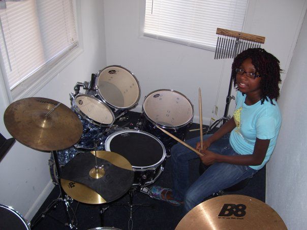 A woman sits in front of a drum set with a cymbal that says b8 on it