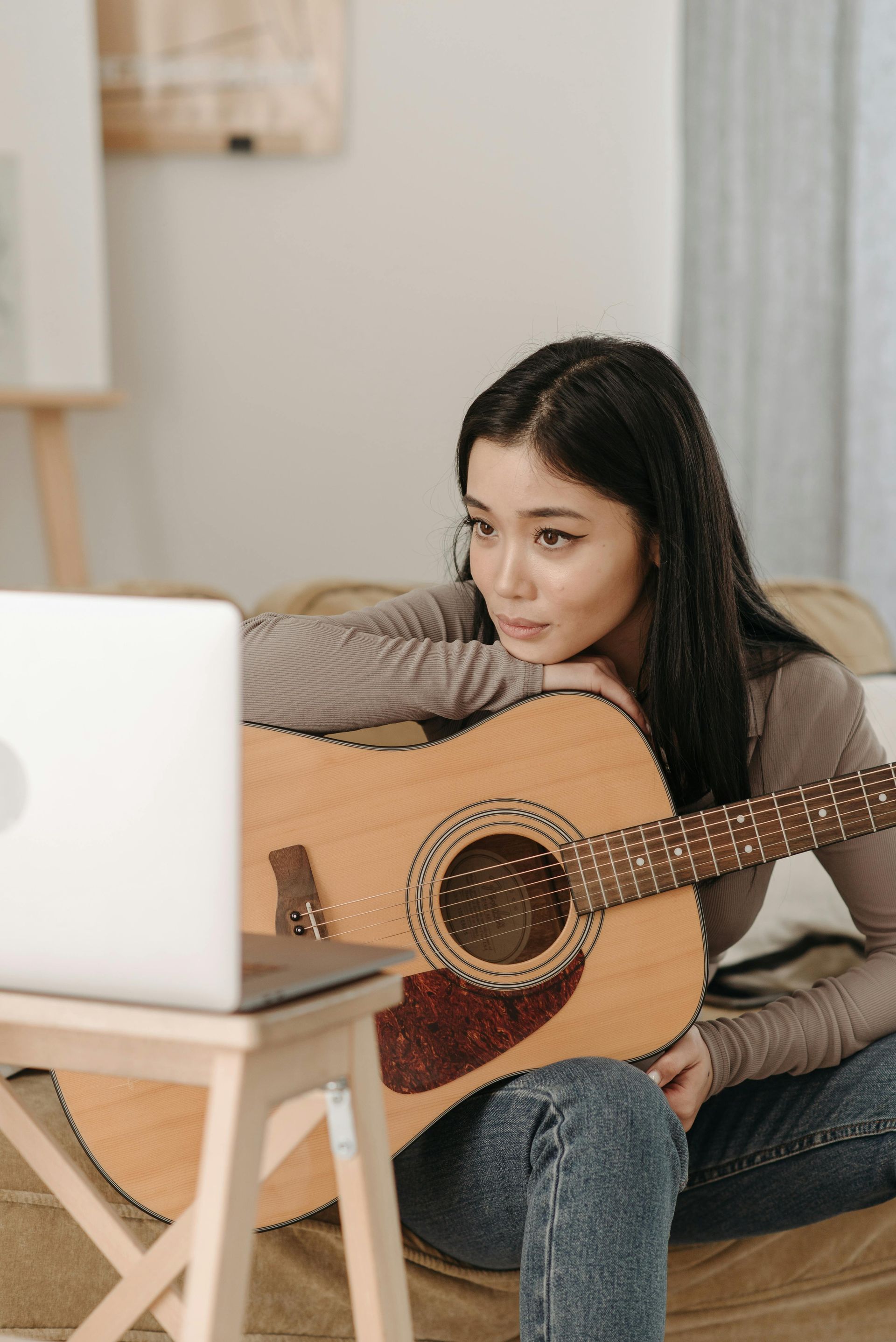 A woman is sitting on the floor holding an acoustic guitar and looking at a laptop.