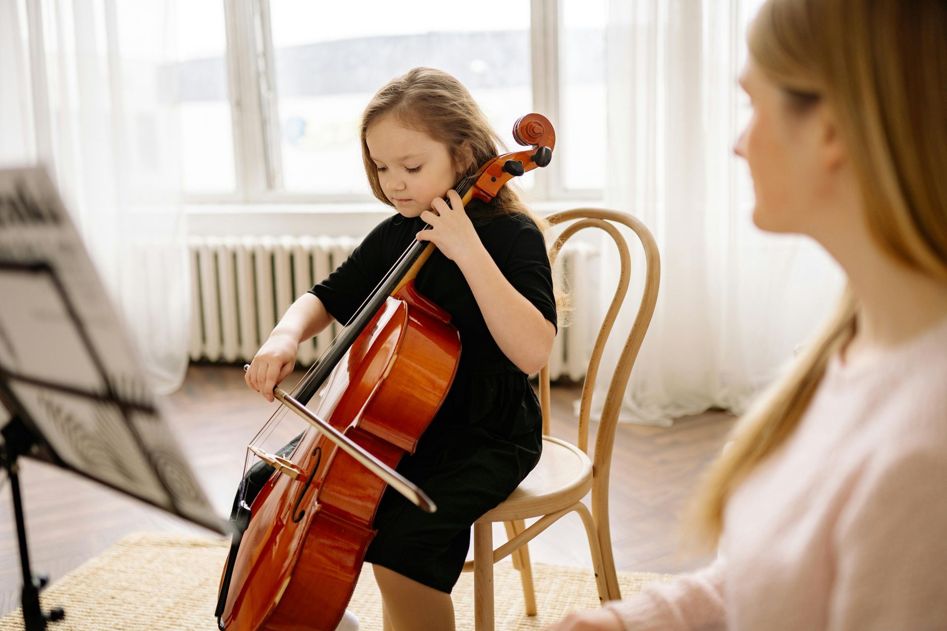 A little girl is playing a cello in front of a woman.