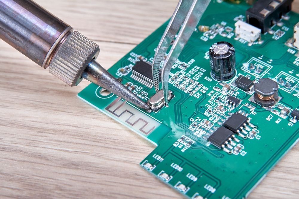 Soldering iron and tweezers working on a green circuit board with small components.