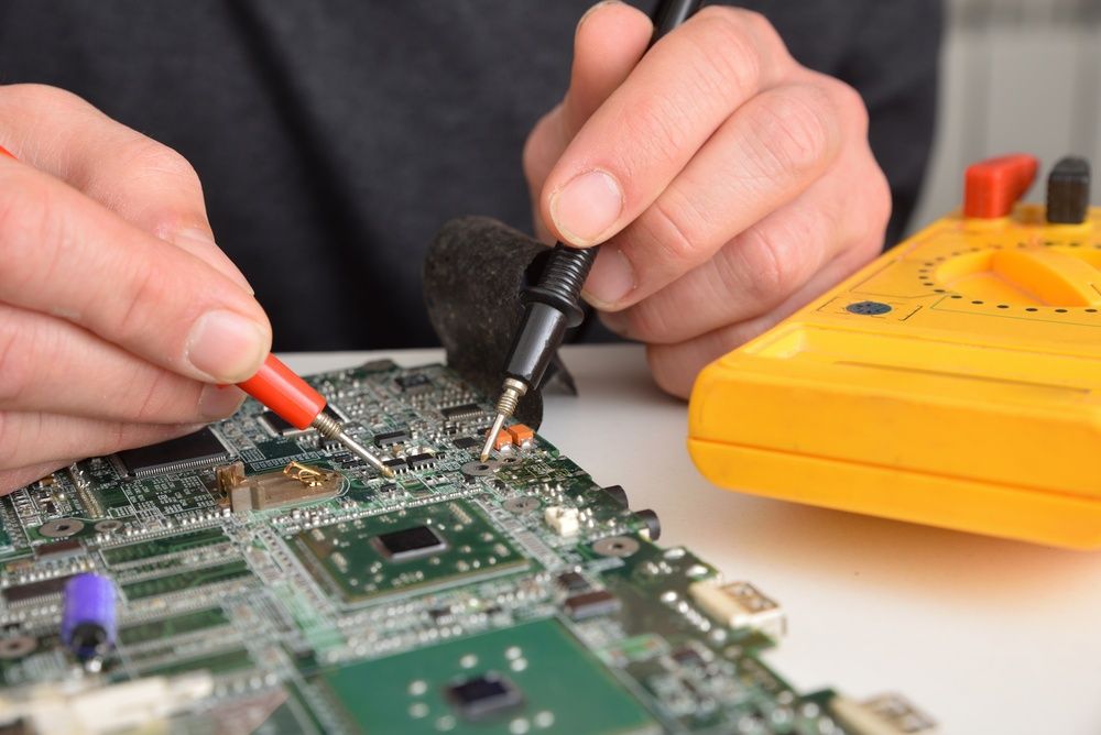 Hands using probes to test a green circuit board beside a yellow multimeter