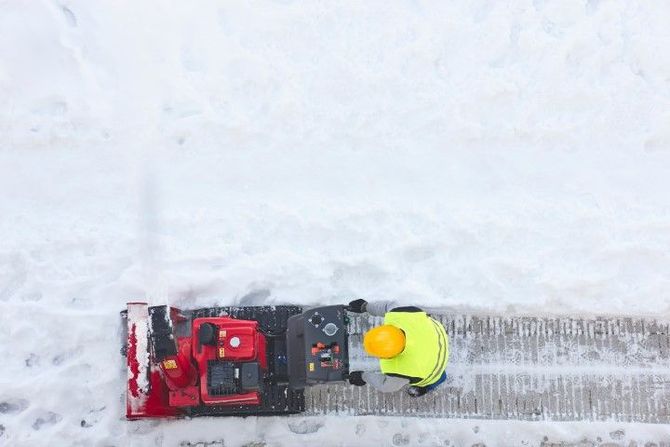 Overhead view of a snowblower clearing a snowy path, with a person in a neon jacket beside it