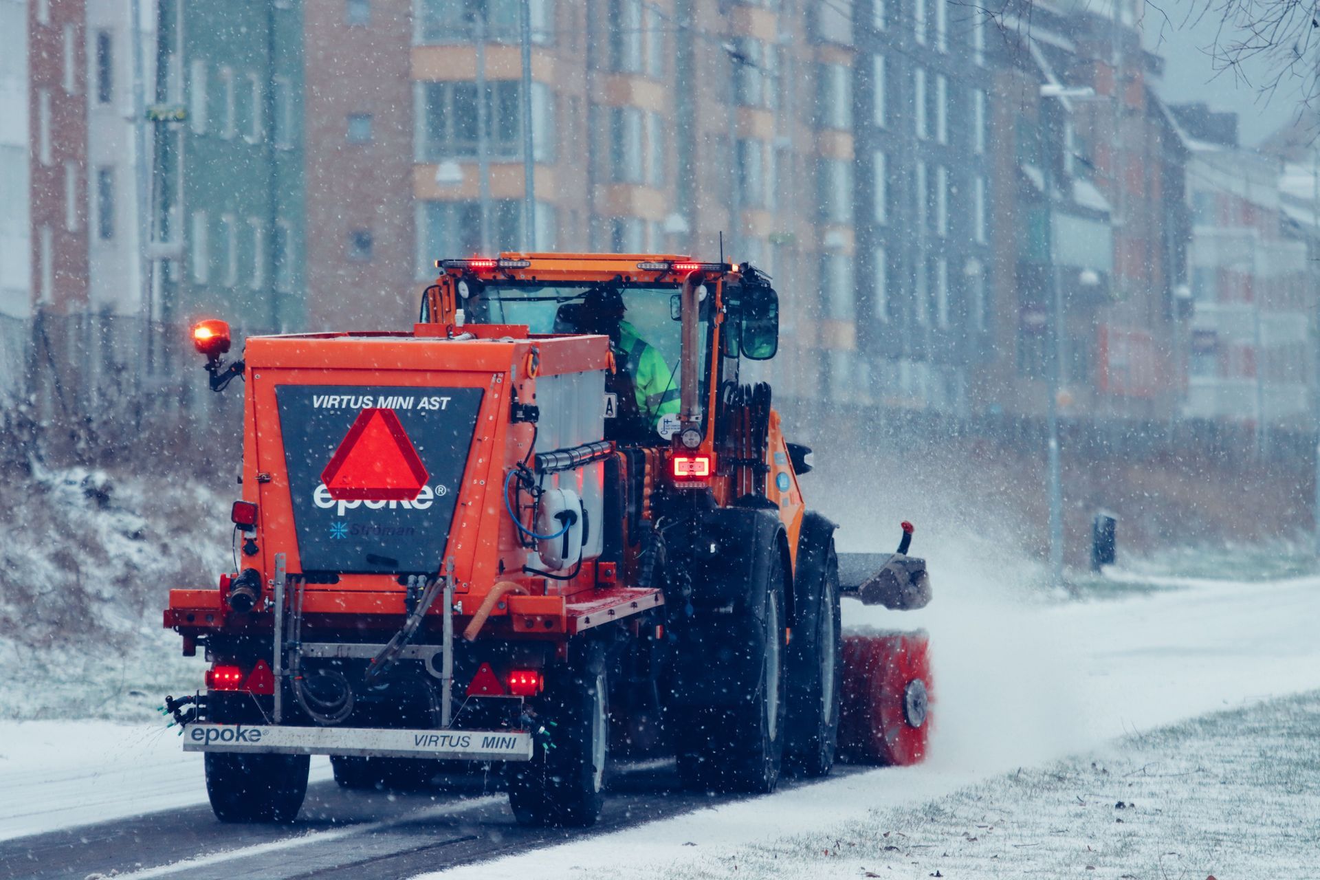Orange snowplow clearing a snowy city street during a heavy snowfall