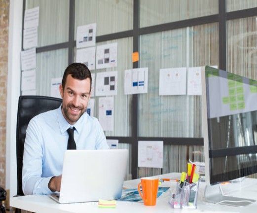 Man at a desk smiles, working on laptop, with notes on the wall behind him. Bright office setting. Man at a desk smiles, working on laptop, with notes on the wall behind him. Bright office setting.