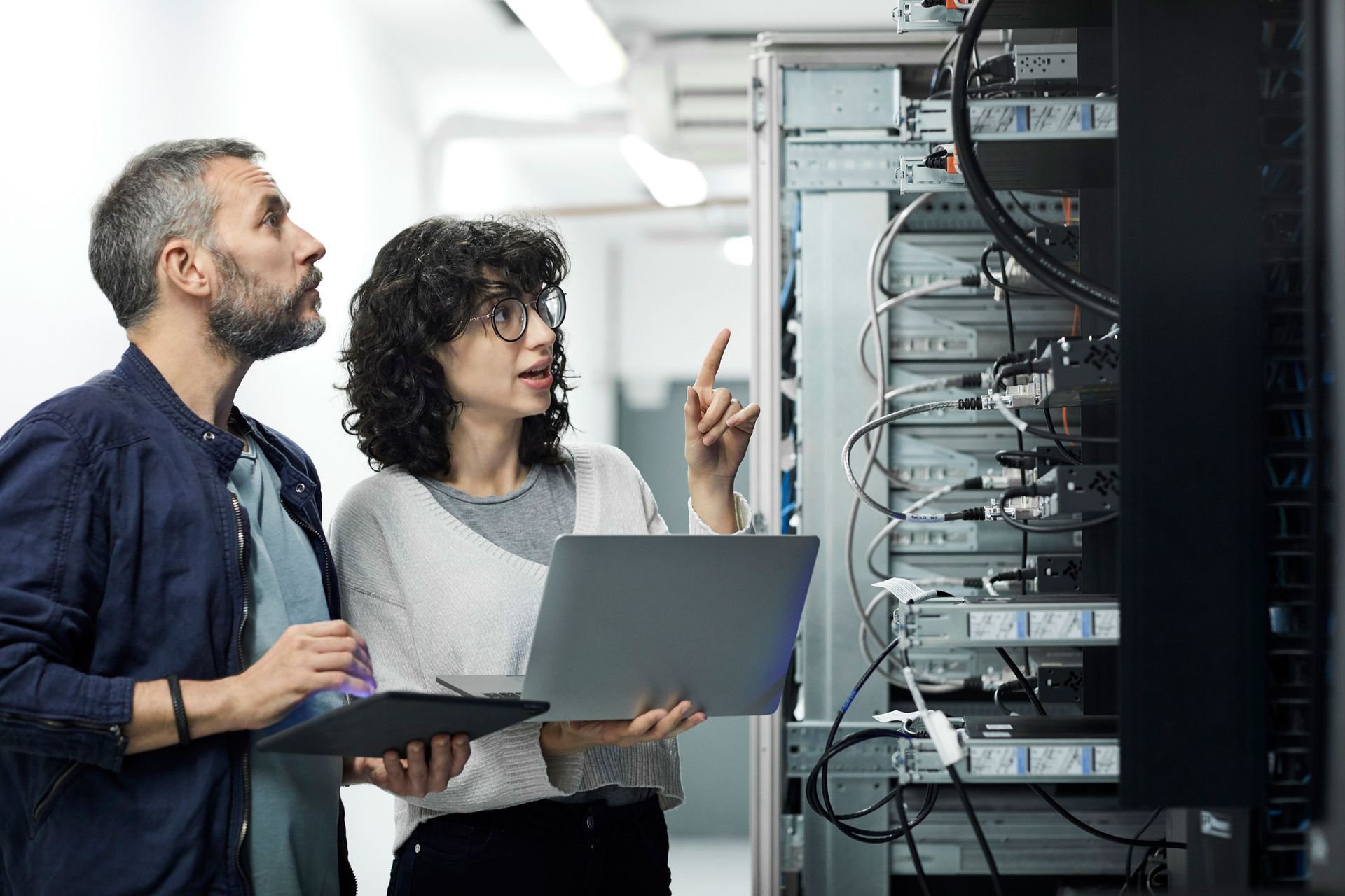 A female technician guiding a male engineer with a digital tablet in a server room. A female technician guiding a male engineer with a digital tablet in a server room.