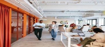 Two people walking in an office with open workspace. The office has white desks, orange curtains, and many people are present.