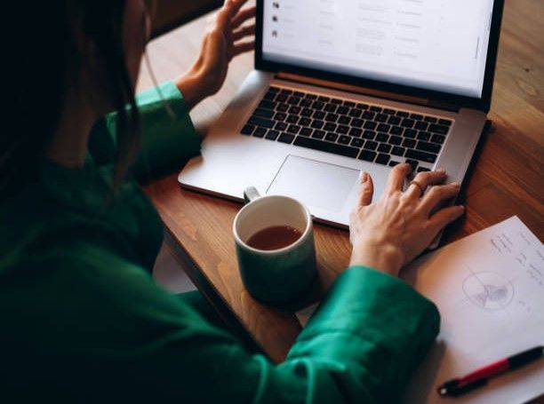Person working on a laptop at a desk, with a mug of tea and notes. Wearing a green top. Person working on a laptop at a desk, with a mug of tea and notes. Wearing a green top.