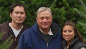 Three people, two women and a man, stand in front of Christmas trees.