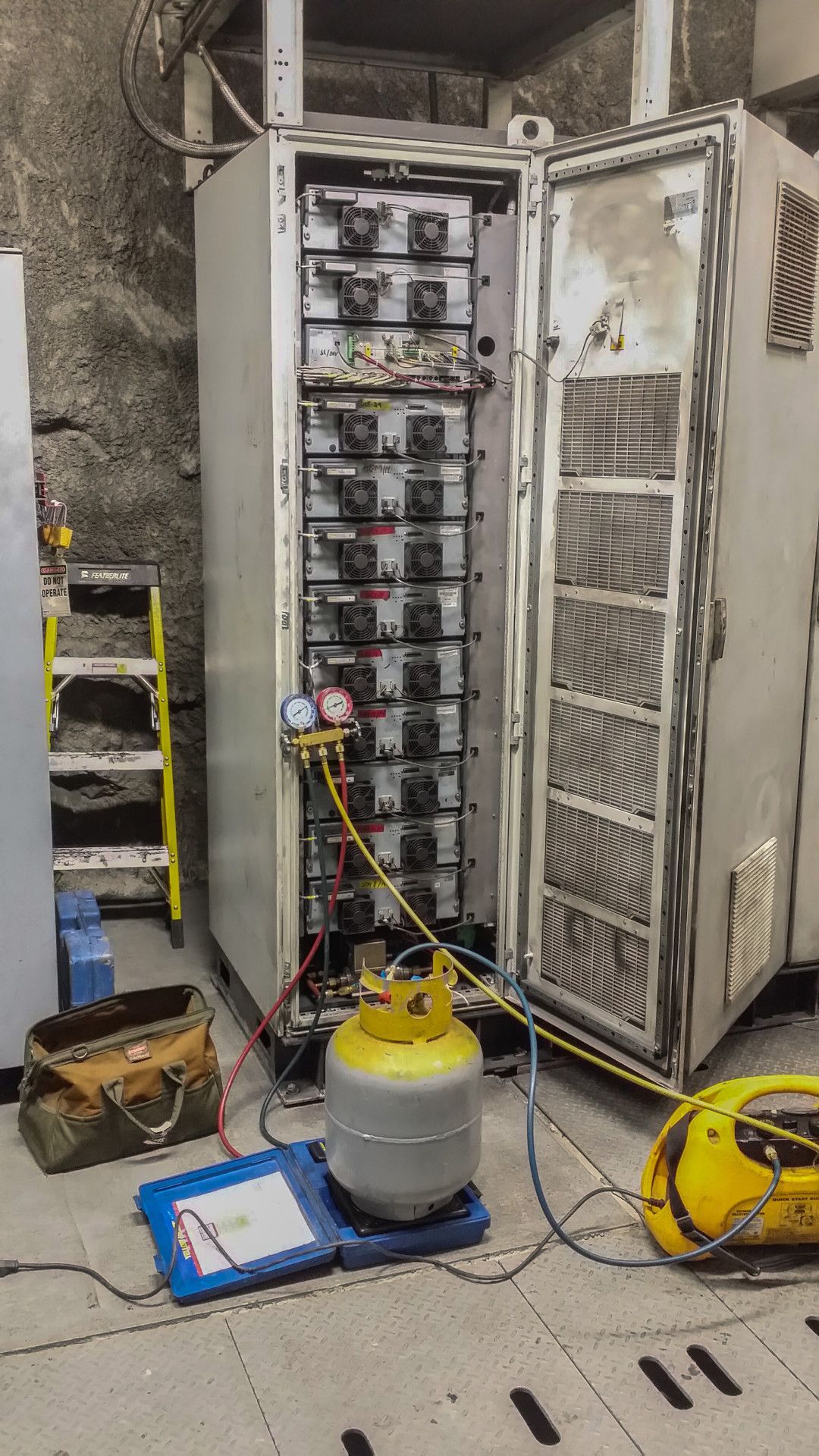 Technician servicing server cabinet with open door, refrigerant tank, and gauges in a utility room.