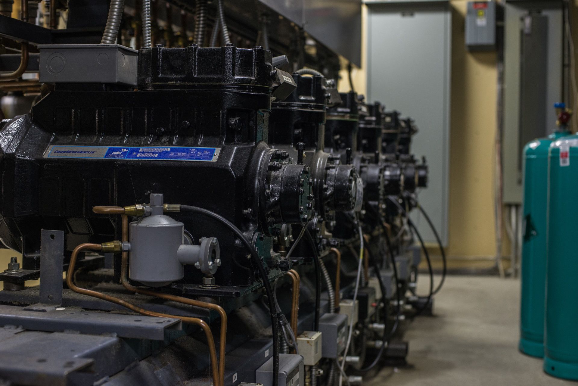 Row of large black industrial compressors in a utility room, copper piping, and a green gas cylinder.