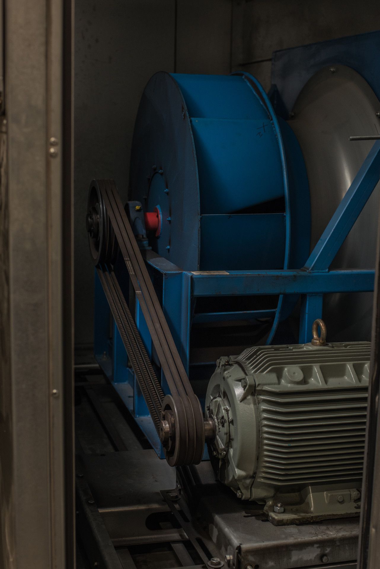 Blue industrial blower fan with a belt connected to a green electric motor, inside a metal enclosure.