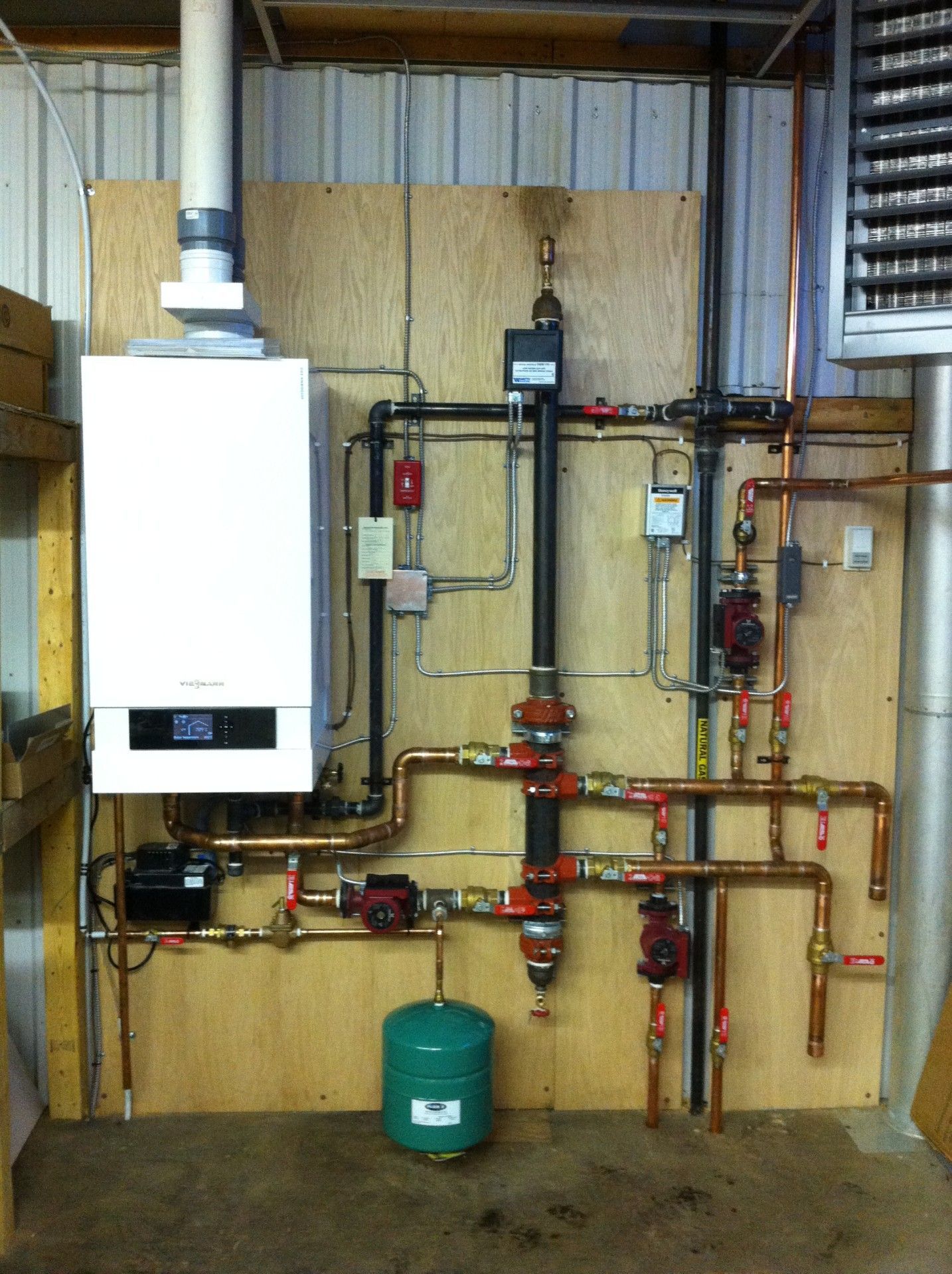 Heating system with white boiler, copper pipes, and green expansion tank mounted on plywood.