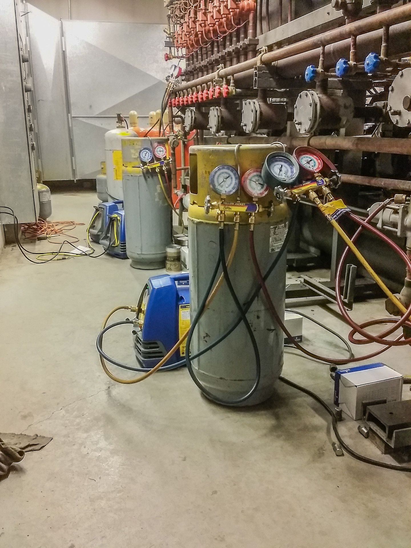 HVAC equipment setup in a utility room. Cylinders, gauges, and hoses connected to a system with pipework in the background.
