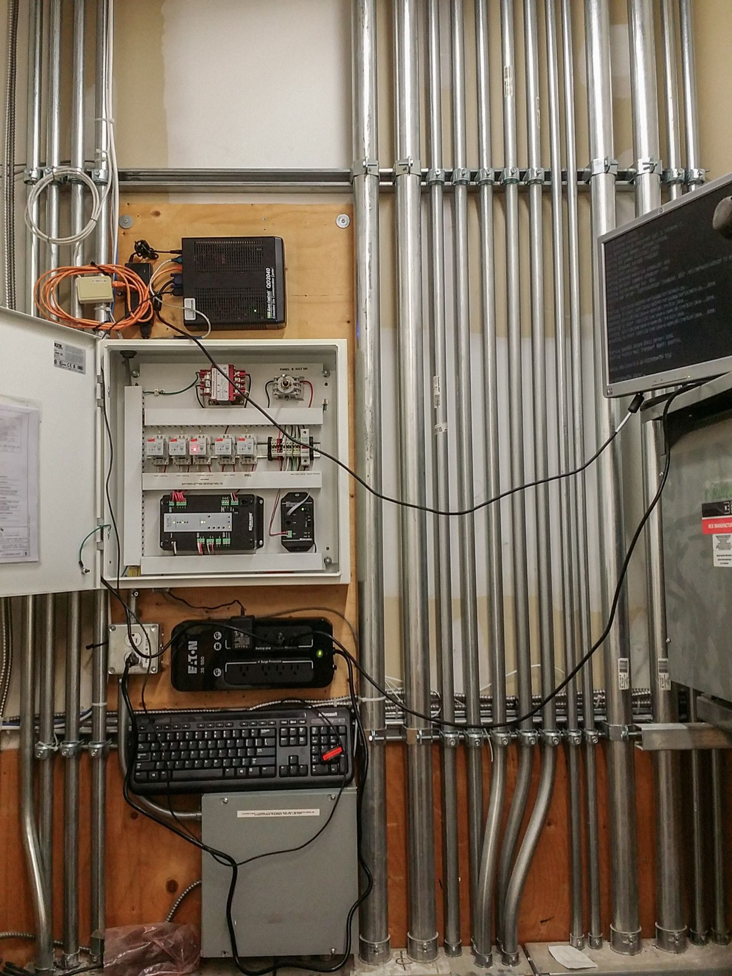 Electrical conduit and control panel mounted on a wood wall; includes a keyboard, monitor, and other components.