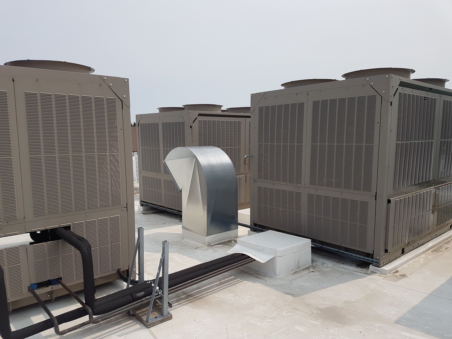 Rooftop view of multiple large HVAC units with a silver ventilation duct.